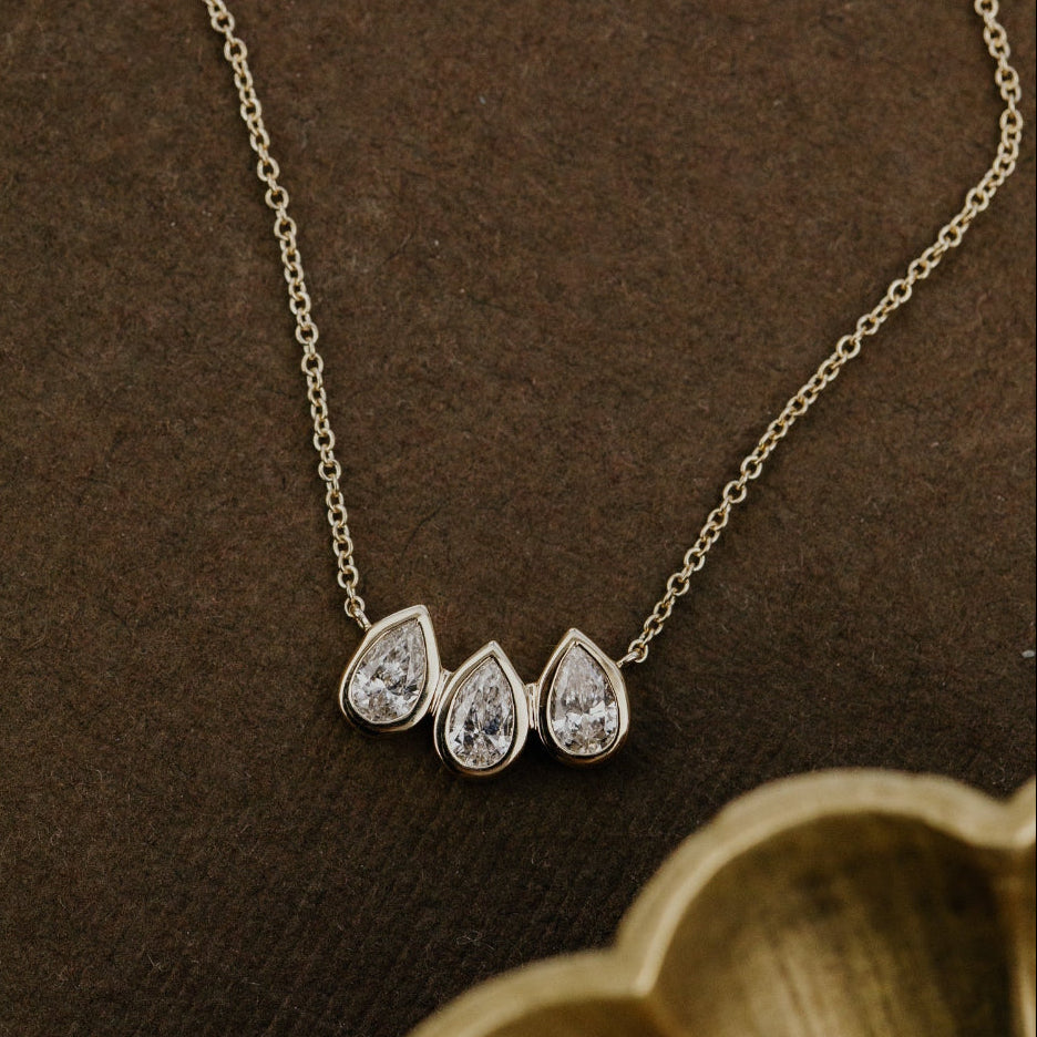 Gold necklace with three pear-shaped lab grown diamonds on a brown background.