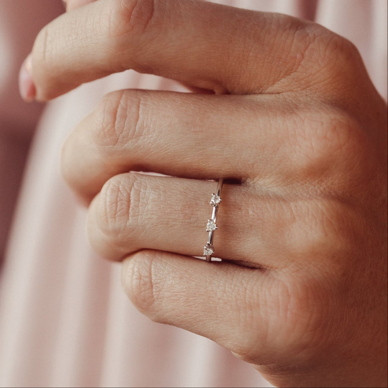 Close-up of a hand wearing a white gold wedding band with three diamonds with a pink background.