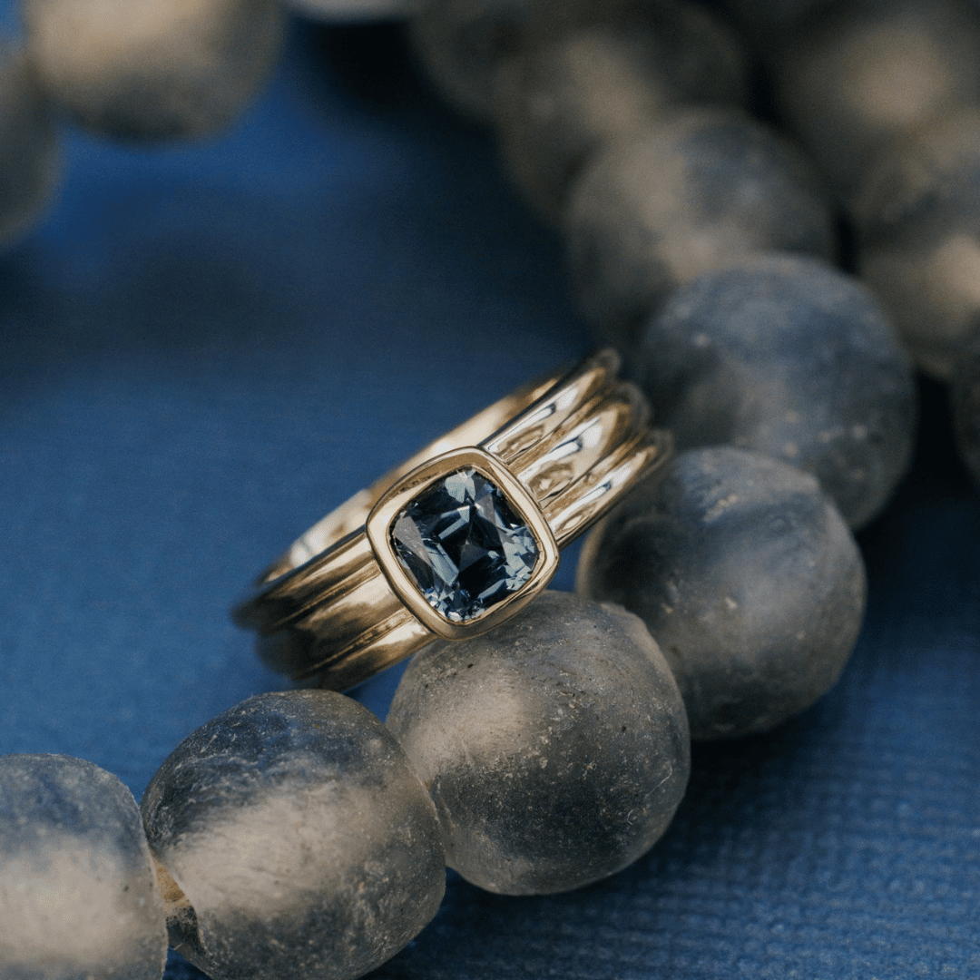 A 14k gold ring featuring a solitare Montana blue sapphire gemstone, displayed on a textured blue surface on a beaded necklace. 