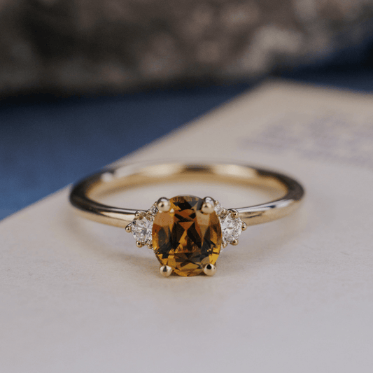 A 14k gold ring featuring an orange-green sapphire gemstone, displayed from the front view on a textured white surface with a soft background.