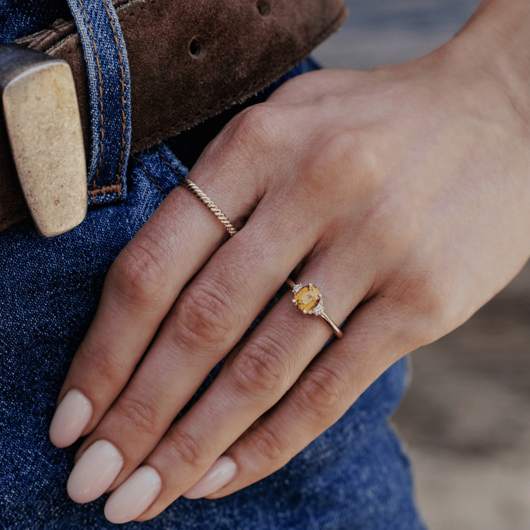 A 14k gold ring featuring an orange-green sapphire gemstone, displayed on the ring finger of a hand model.