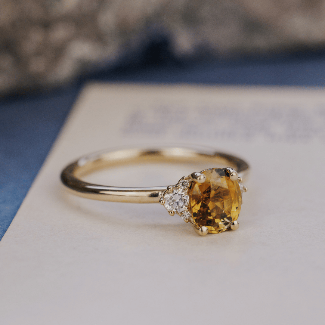 A 14k gold ring featuring an orange-green sapphire gemstone, displayed from a side view on a textured white surface with a soft background.