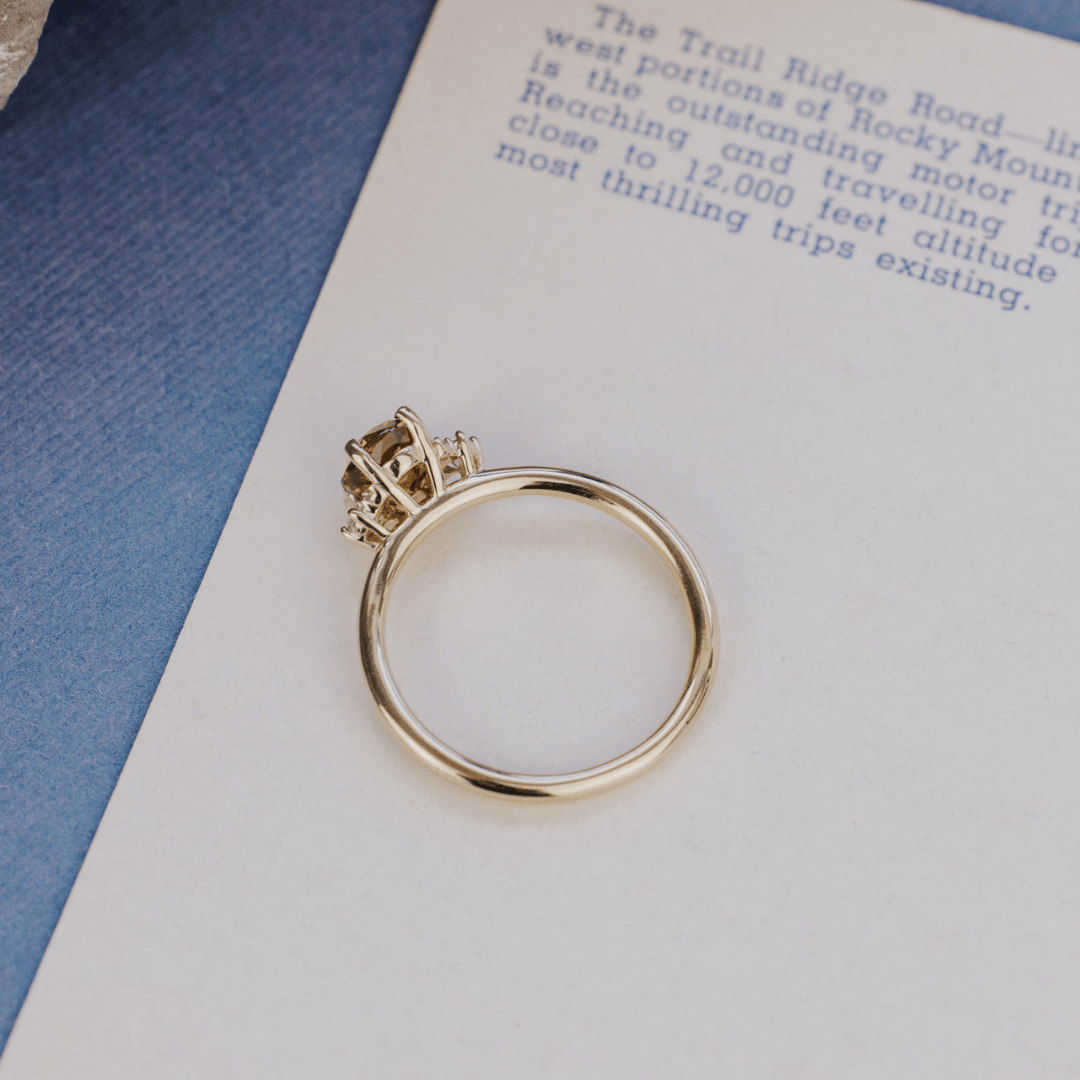 A 14k gold ring featuring an orange-green sapphire gemstone, displayed from a top view on a textured white surface.