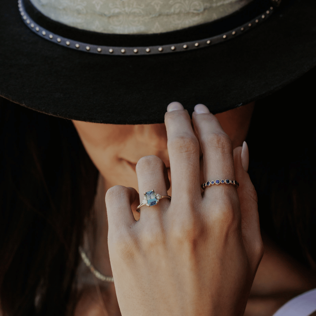 A 14k gold ring featuring a blue sapphire gemstone, on a hand model tipping her hat.