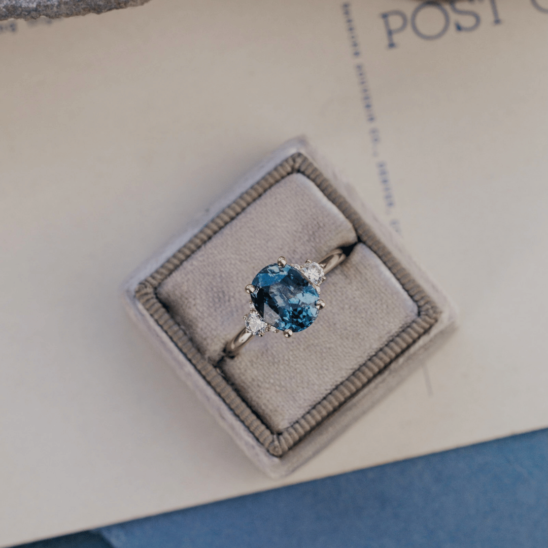 A 14k gold ring featuring a blue sapphire gemstone viewed from a ring box, displayed on a textured white surface.