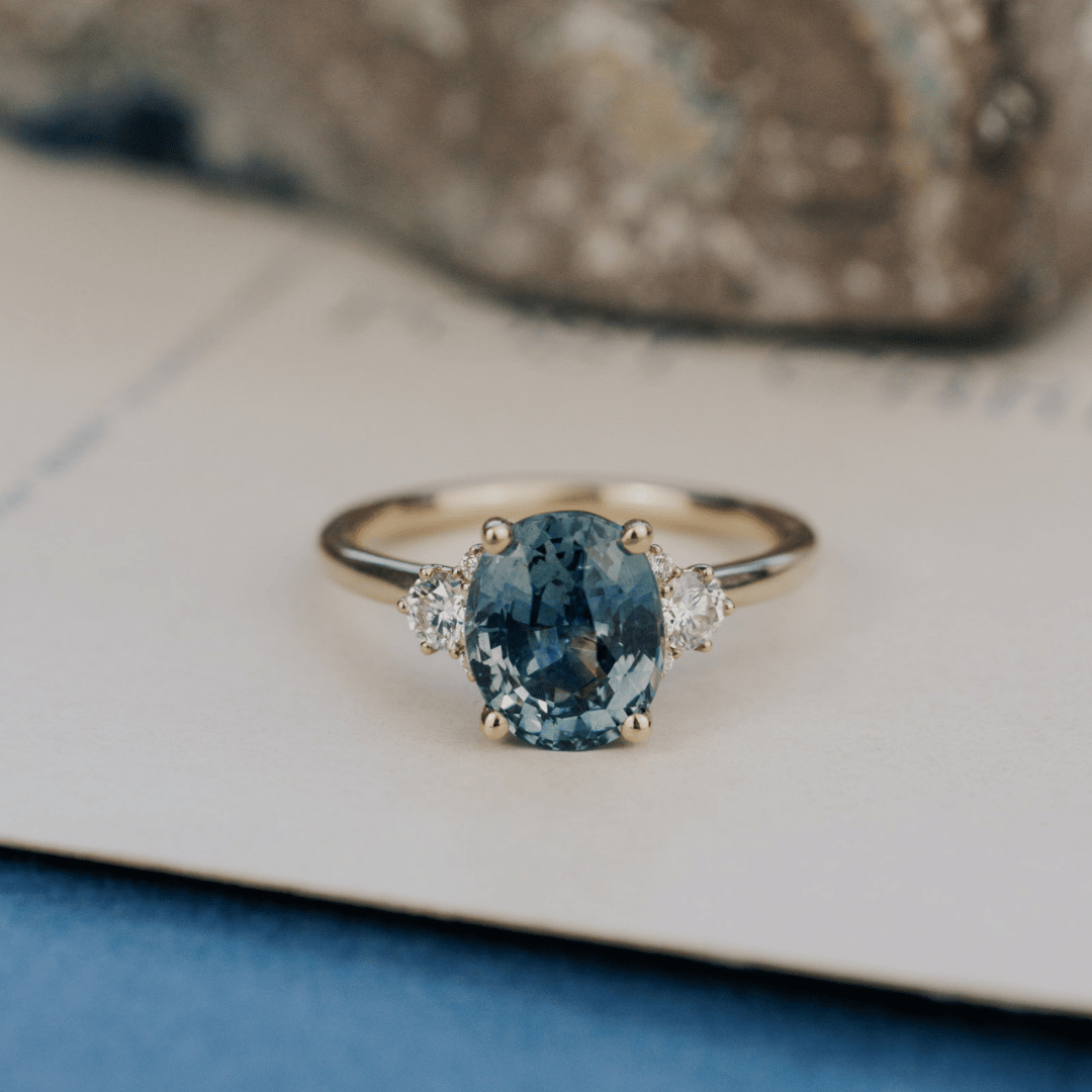 A 14k gold ring featuring a blue sapphire gemstone viewed from straight on, displayed on a textured white surface with a soft-focus background.