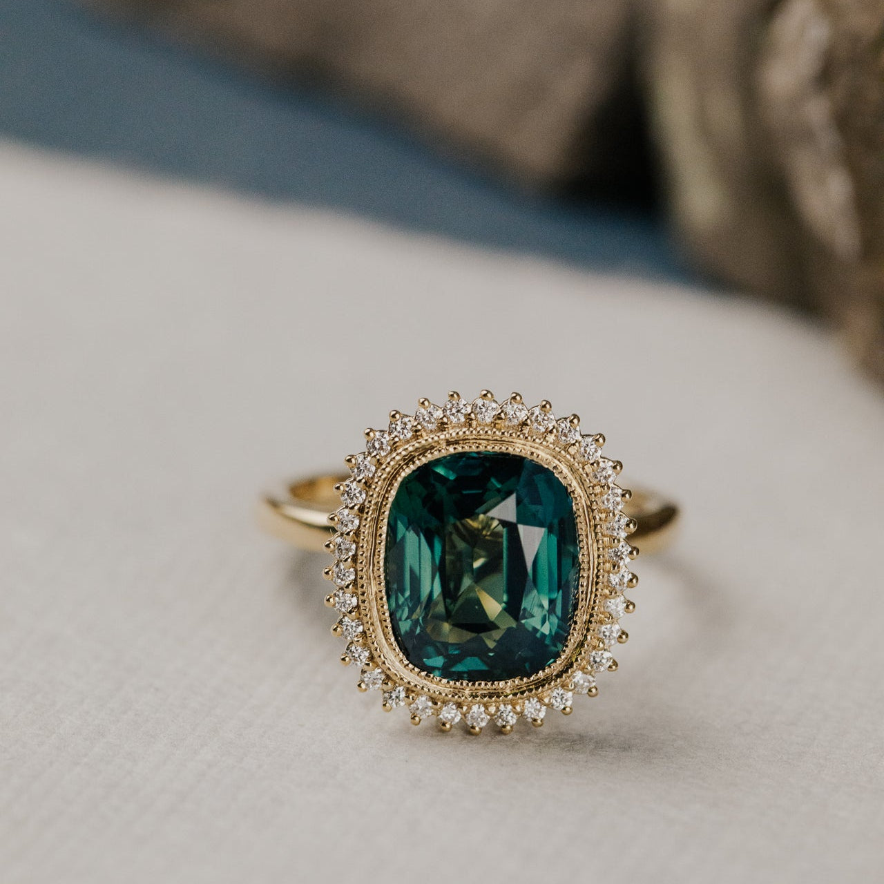 Gold diamond halo ring with a elongated cushion cut green Madagascar sapphire center stone on a textured surface