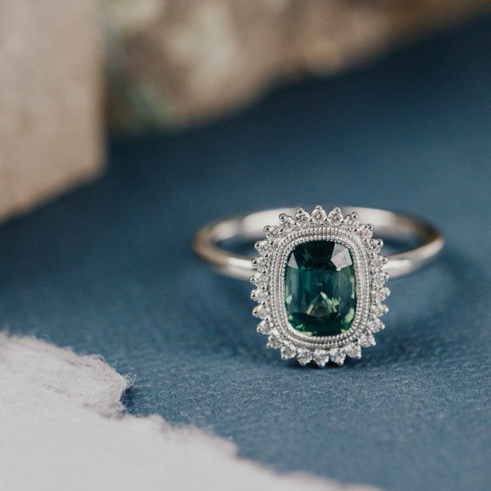 White gold engagement ring with a teal sapphire center stone and a diamond halo on a blue surface