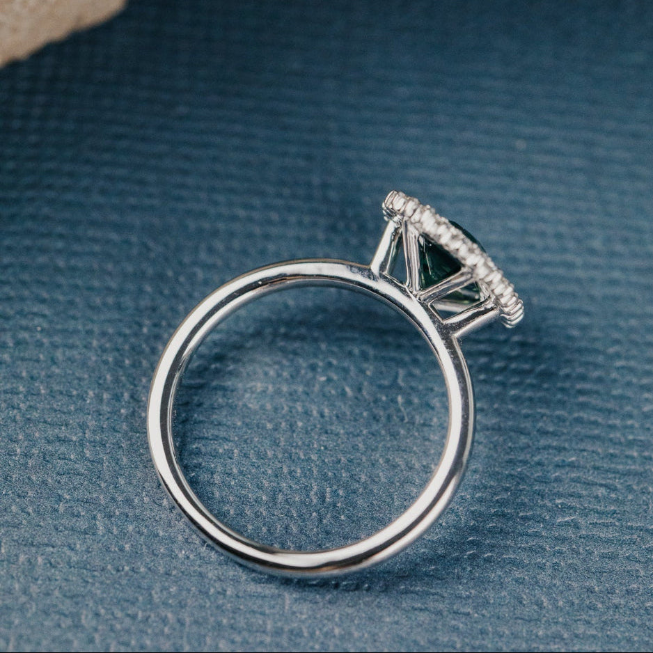 Top-down view of a white gold engagement ring with a teal sapphire center stone and a diamond halo on a textured blue surface