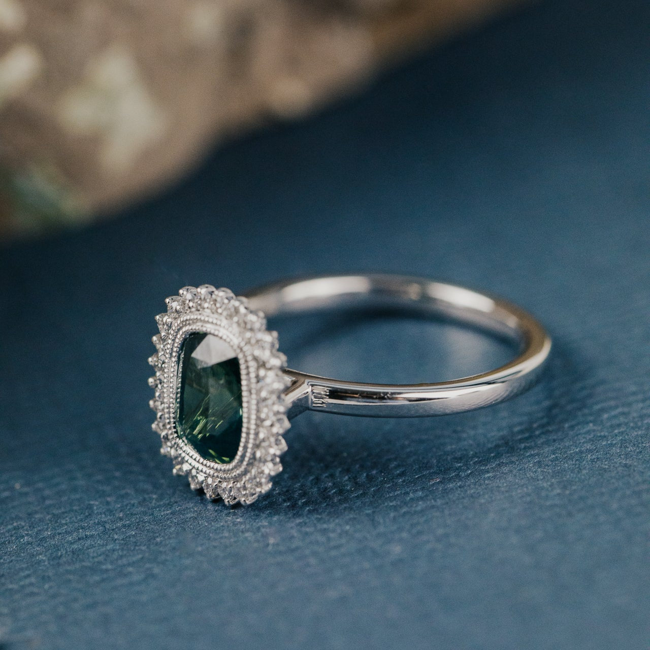 Side profile of a white gold engagement ring with a teal sapphire center stone and a diamond halo on a blue fabric background