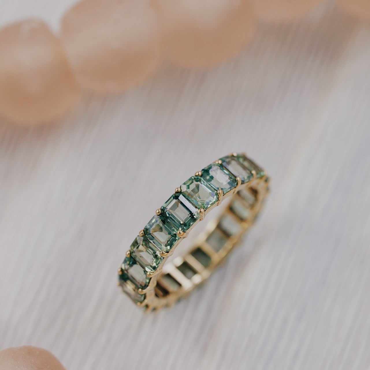 The Amara 5.55 cttw Green Emerald Cut Sapphire Eternity Band in 14k yellow gold, featuring emerald cut green natural sapphires set in prongs in a single row eternity design, shown at an angle on a light textured surface.
