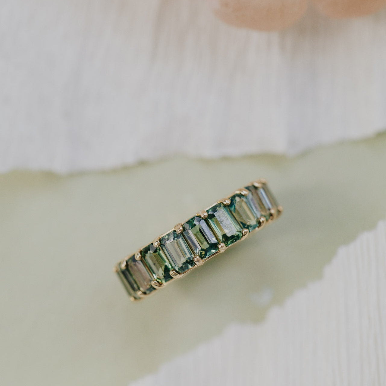 Gold Amara band with green sapphires on a light fabric background.