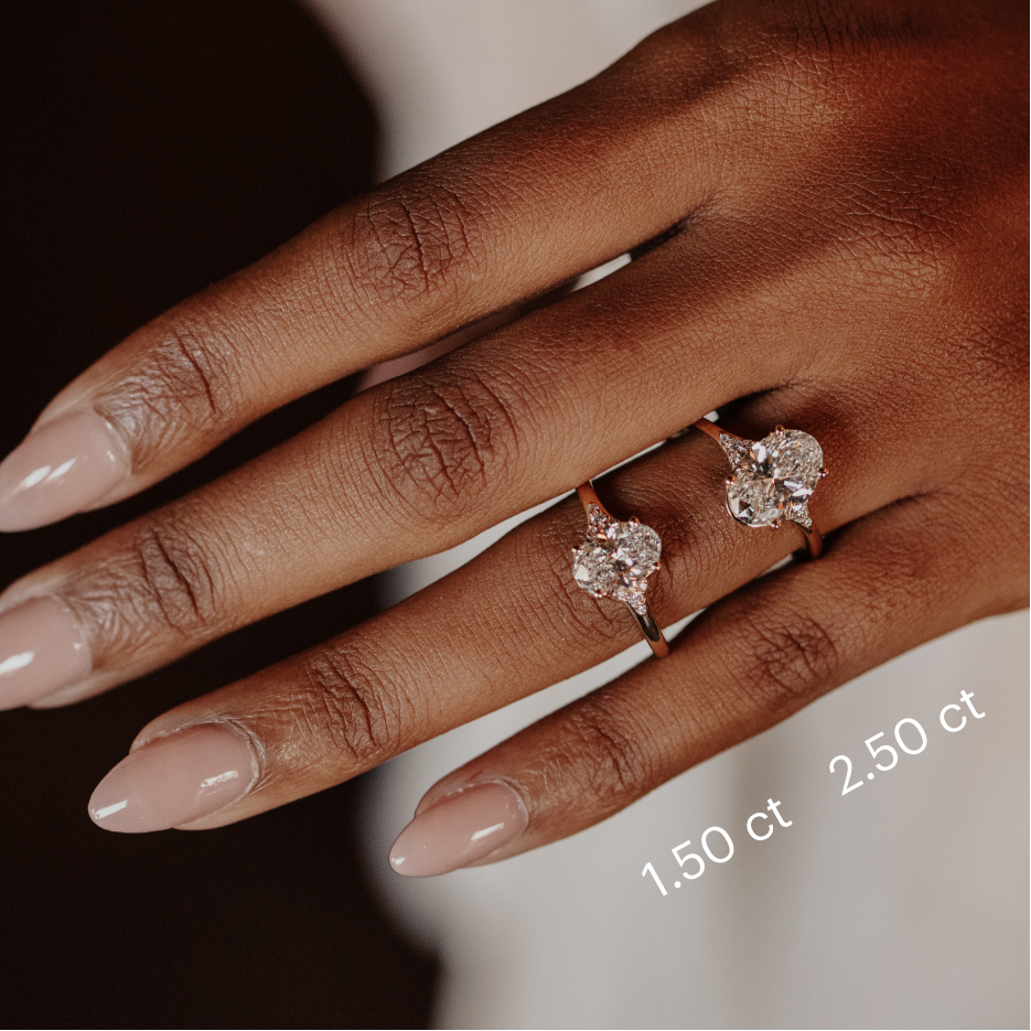 Close up of a hand wearing two Angie oval engagement rings in different carat weights with a size chart. 