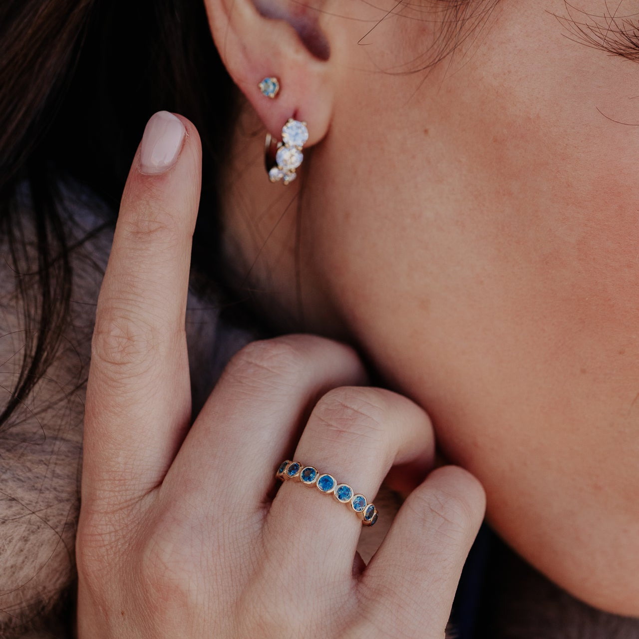 Close up of a person wearing the Aster round Montana sapphire band, Montana sapphire stud earrings, and a three diamond hoop earring.