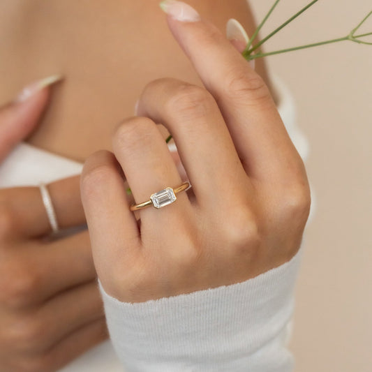 A woman's hand wearing a Austen gold solitaire ring with an emerald cut diamond, set in a half bezel design.