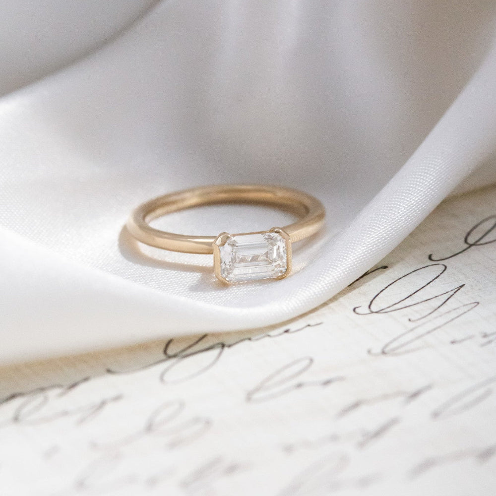 Gold Austen ring with a emerald-cut diamond on a white ribbon and paper background.