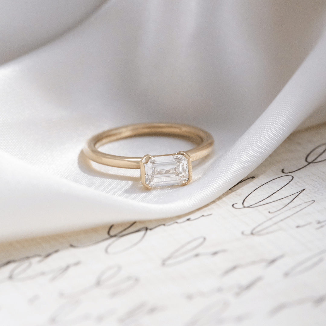 Gold Austen ring with a emerald-cut diamond on a white ribbon and paper background.