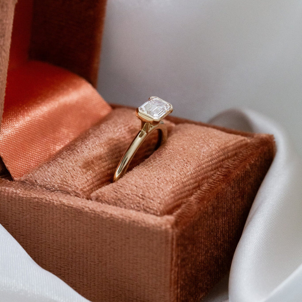 Gold Austen ring with a emerald-cut diamond on a brown cushion in a box.