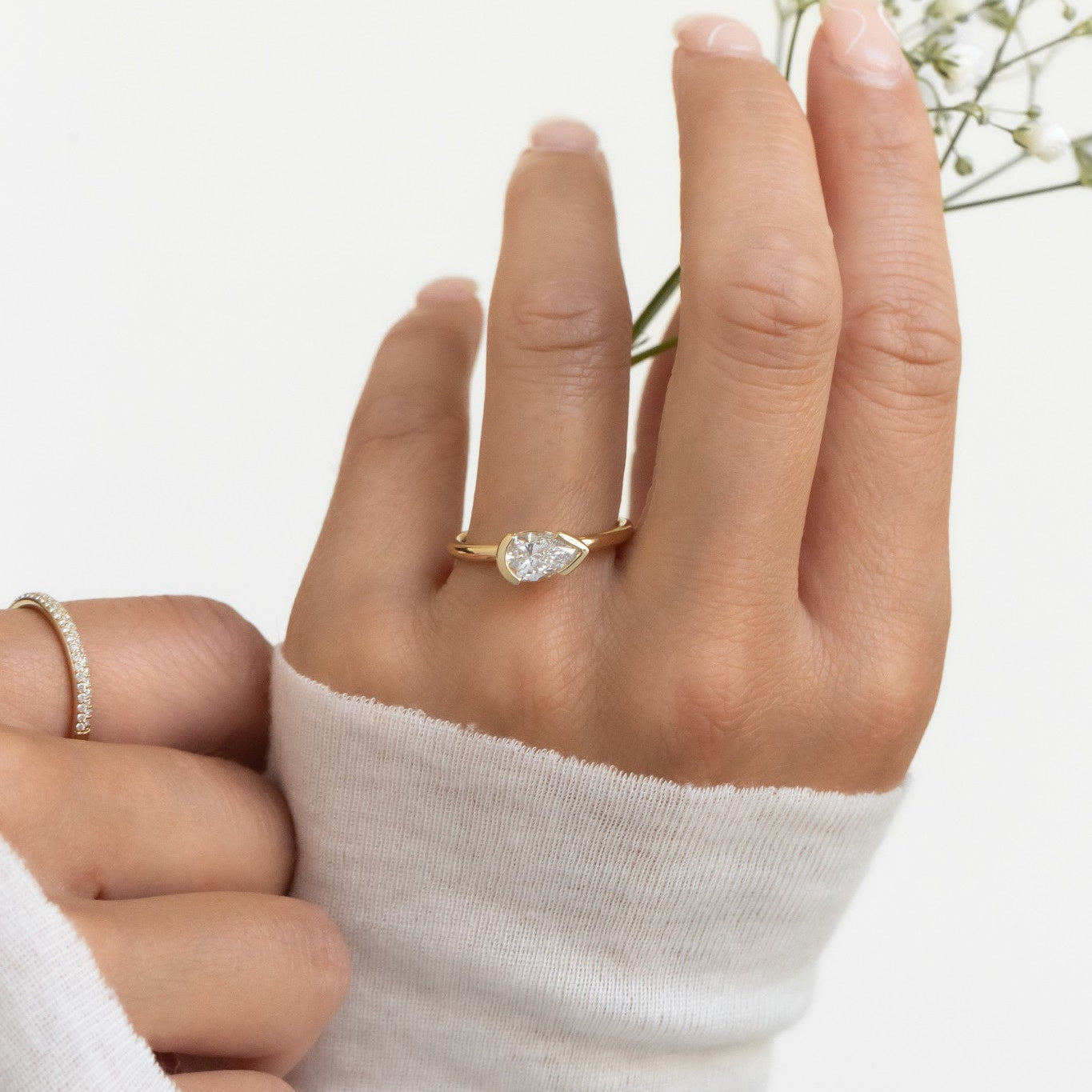 A hand wearing a white gold Austen solitaire ring with a pear-shaped diamond, with a white sleeve and holding a small white flower.
