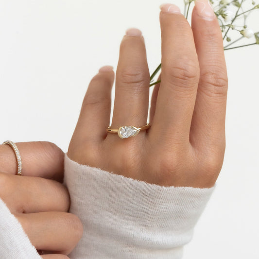 A hand wearing a white gold Austen solitaire ring with a pear-shaped diamond, with a white sleeve and holding a small white flower.