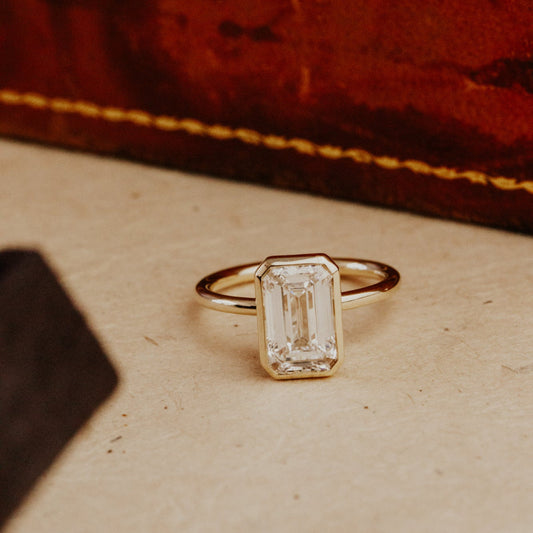 Gold Bailey emerald cut engagement ring with a rectangular diamond on a textured surface.