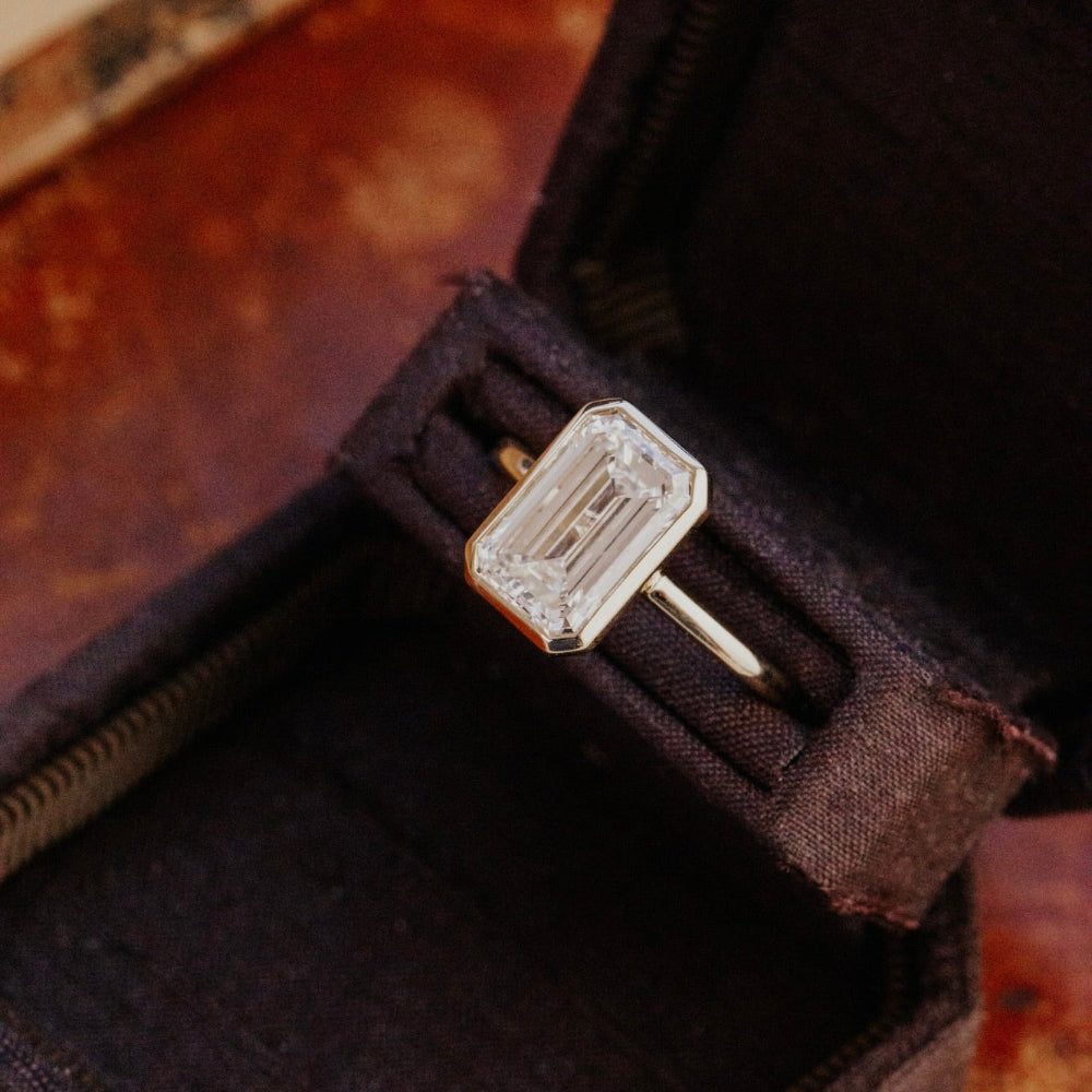 Gold Bailey emerald cut engagement ring with a diamond on a dark fabric background.