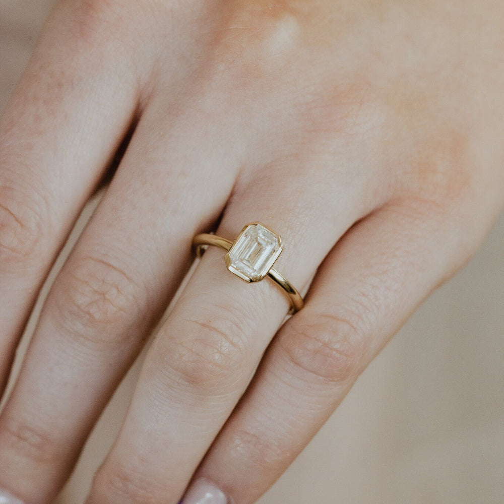 Gold Bailey engagement ring with a emerald cut diamond on a person's finger against a blurred background.
