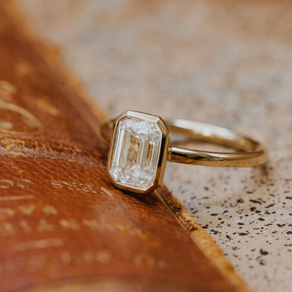 Gold Bailey engagement ring with a emerald cut diamond on a textured surface.
