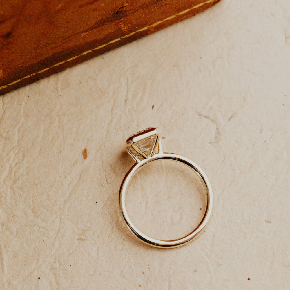 Gold Bailey emerald cut diamond ring on a textured beige surface with a wooden block in the background.