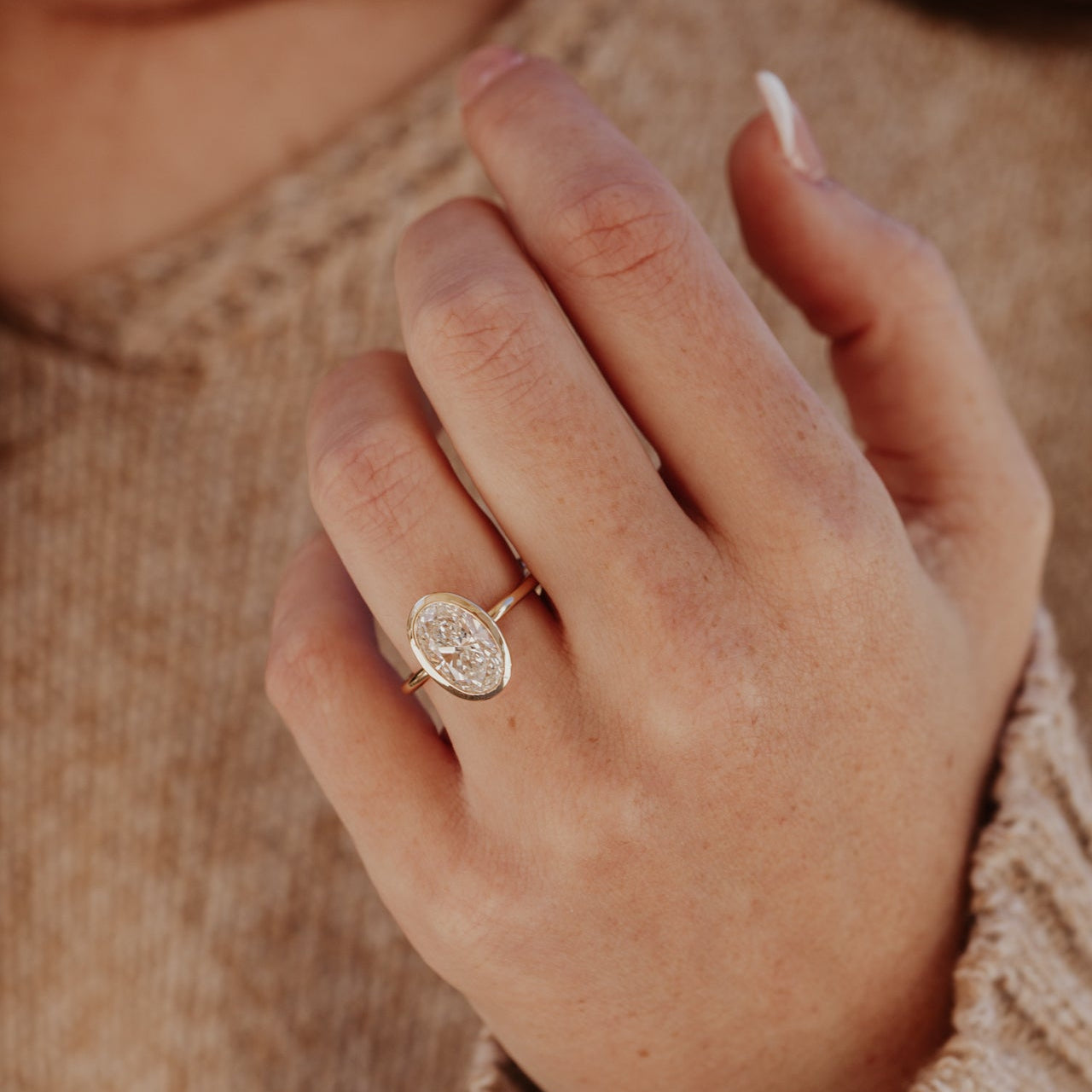 Close-up of a hand wearing a gold Bailey ring with a oval diamond on a blurred background.