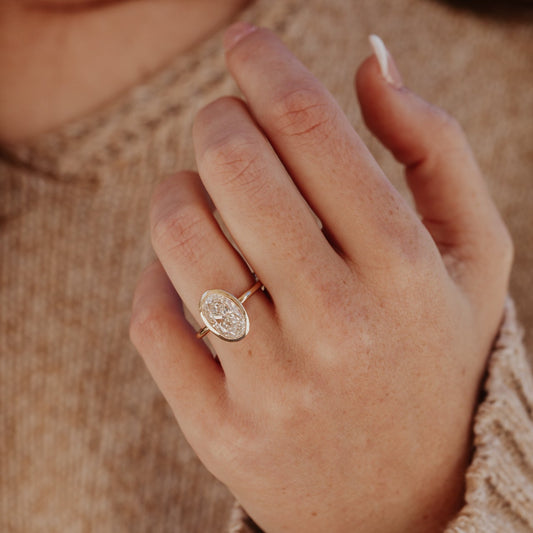 Close-up of a hand wearing a gold Bailey ring with a oval diamond on a blurred background.