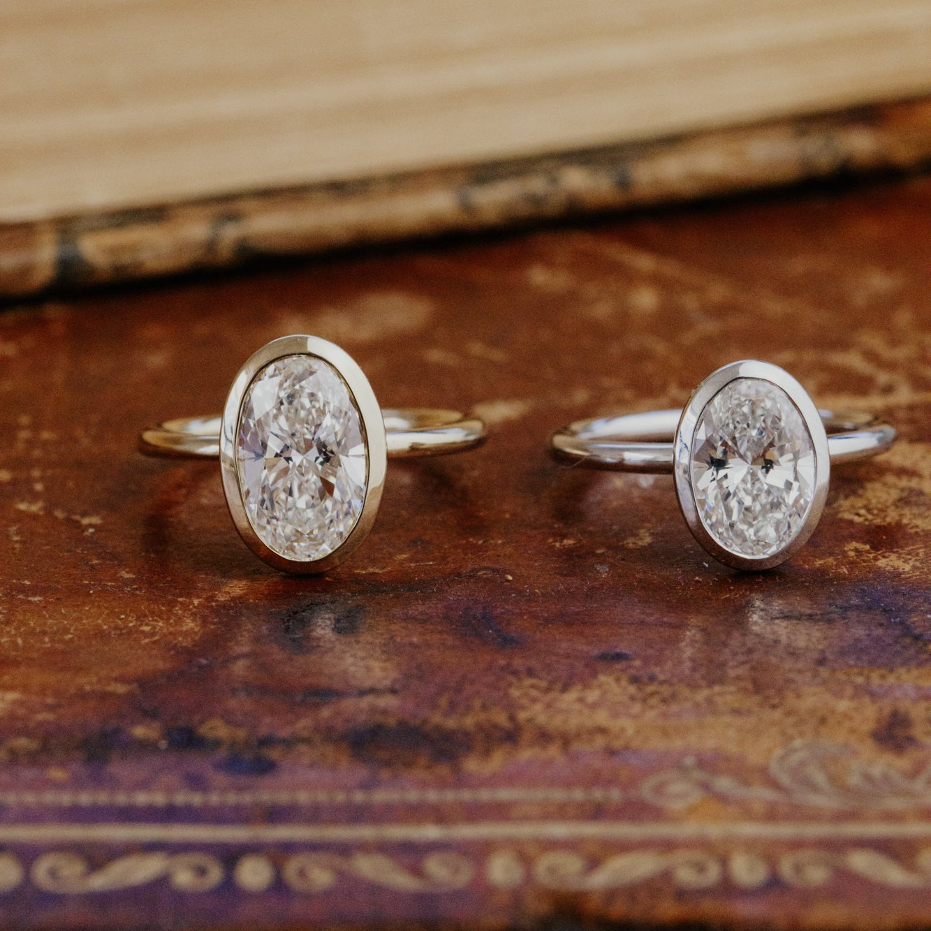 Two Bailey oval diamond rings in different ct weights on a textured wooden surface.