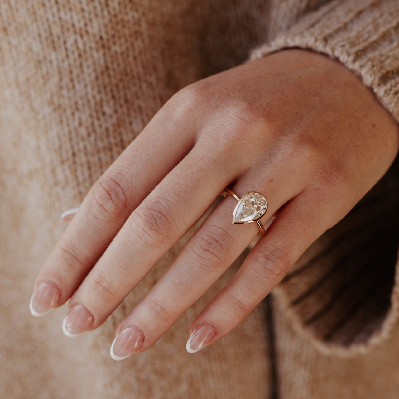 Hand wearing a gold Bailey ring with a pear-shaped diamond on a neutral background.