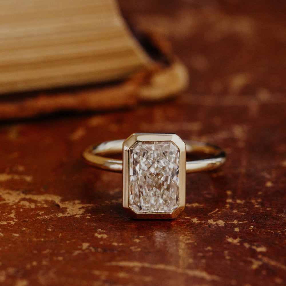Gold Bailey engagement ring with a radiant cut diamond on a textured brown surface.