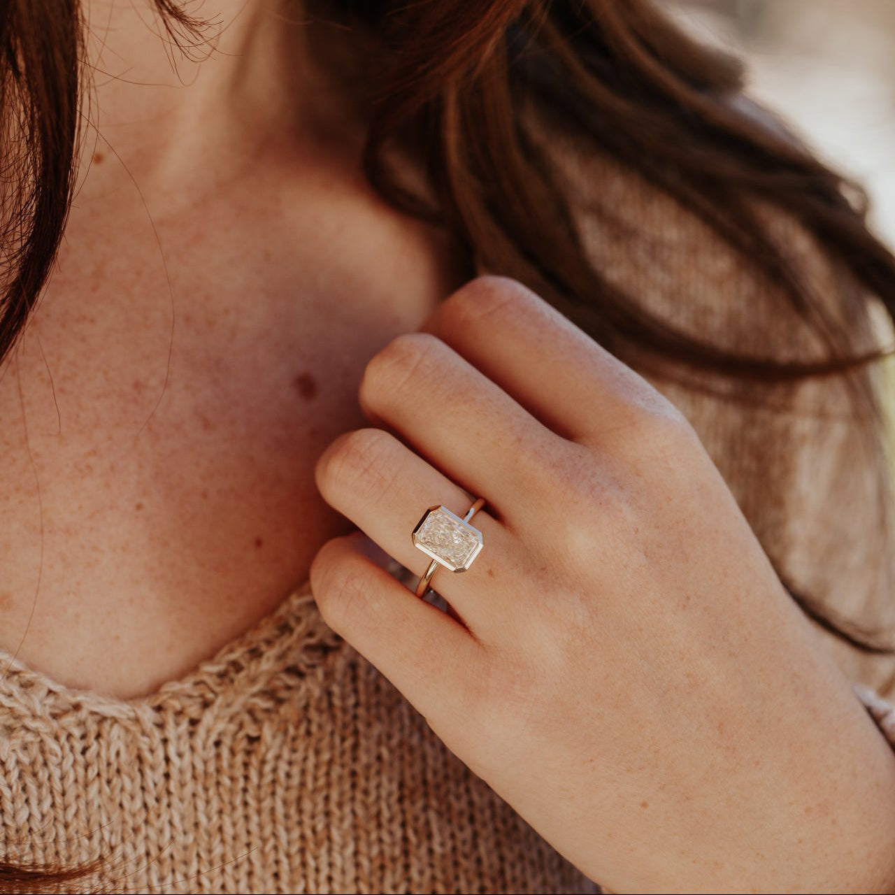 Close-up of a hand wearing a Bailey radiant cut diamond engagement ring, touching the chest.