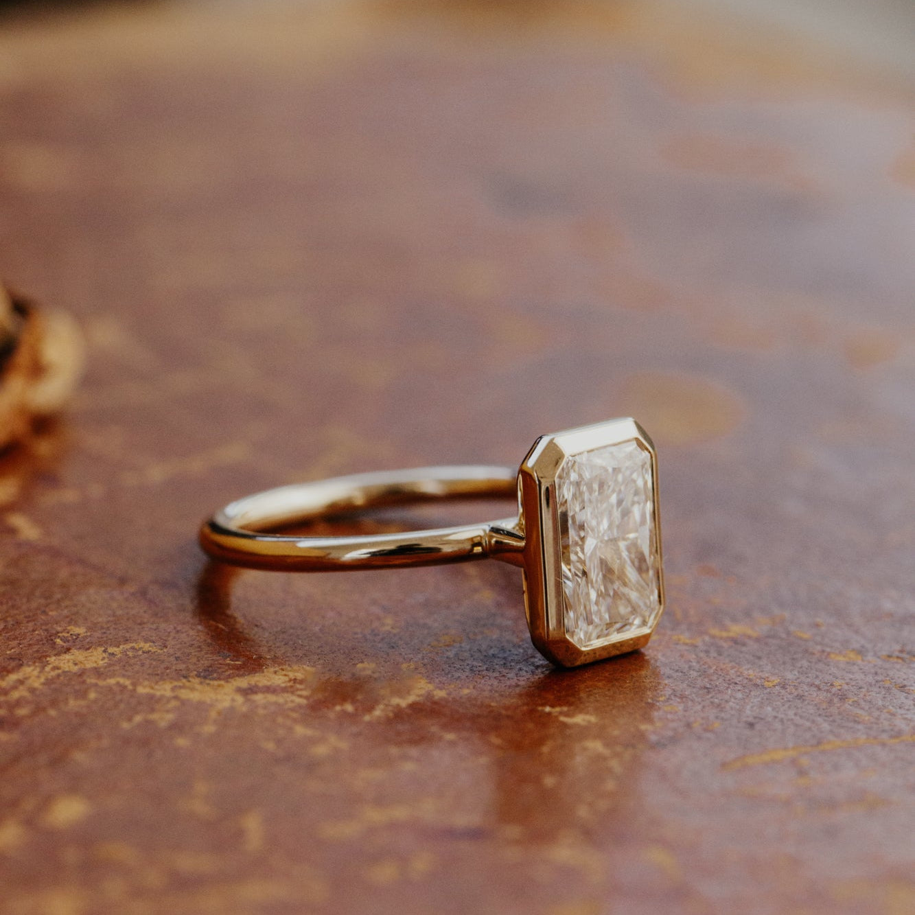 Gold Bailey radiant cut diamond engagement ring with a textured diamond on a brown surface.