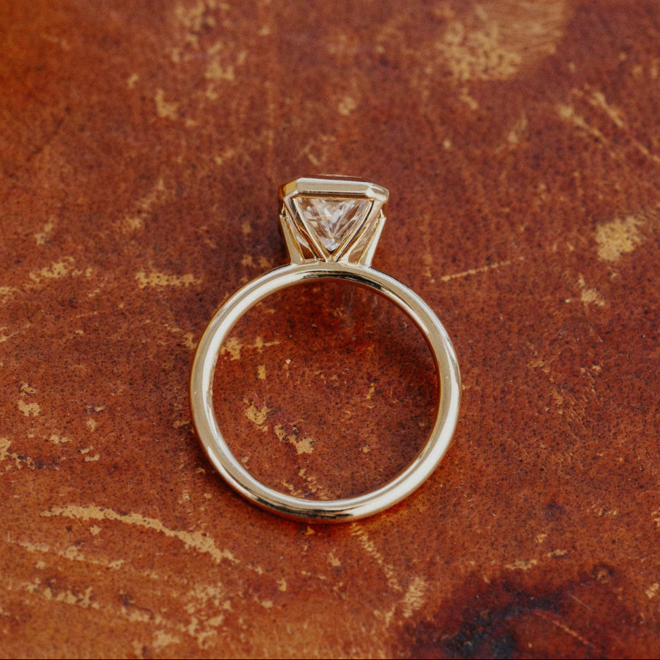 Gold Bailey radiant cut diamond engagement ring on a textured brown surface.