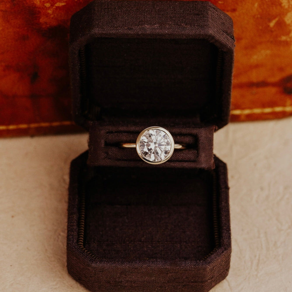 Bailey round diamond solitaire ring in a brown velvet box on a textured surface.