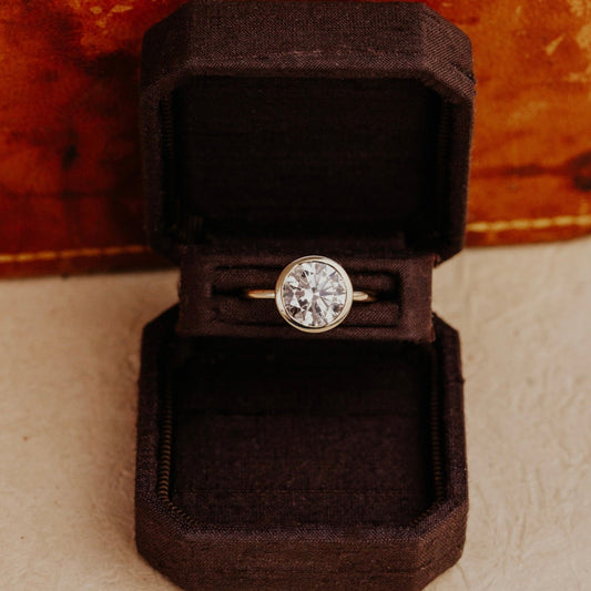 Bailey round diamond solitaire ring in a brown velvet box on a textured surface.
