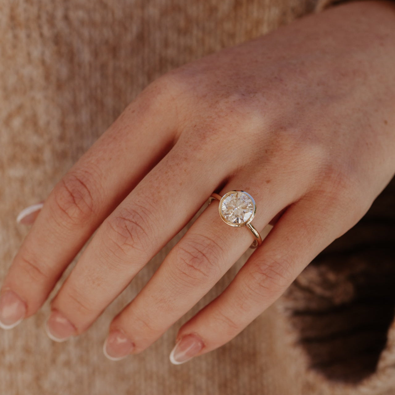 Hand wearing a gold Bailey ring with a round diamond on a textured surface.