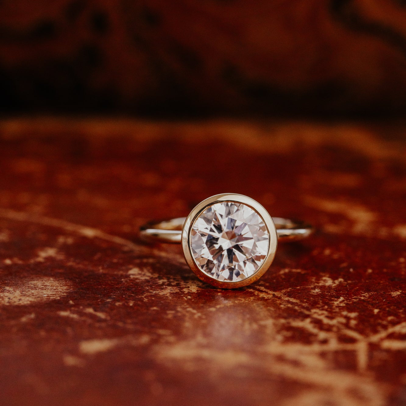 Gold Bailey engagement ring with a large oval diamond on a textured brown surface.