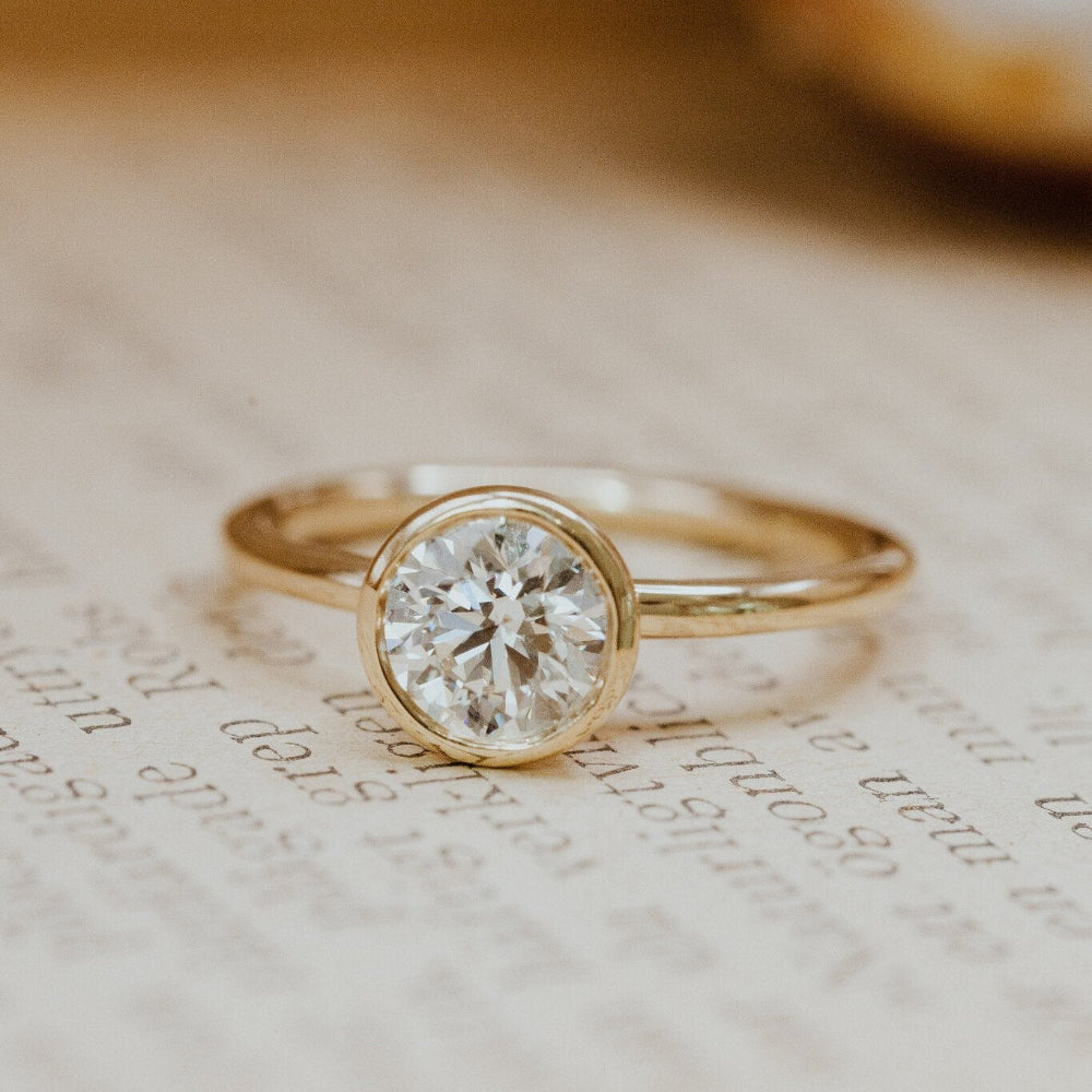 A Bailey round solitaire ring with a bezel setting, featuring a white diamond, placed on a written document background.