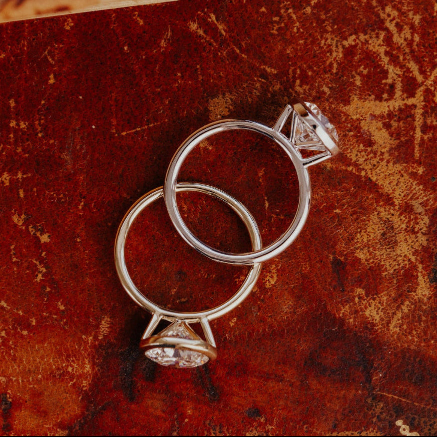 Two white gold Bailey engagement rings with round diamonds on a textured brown surface.