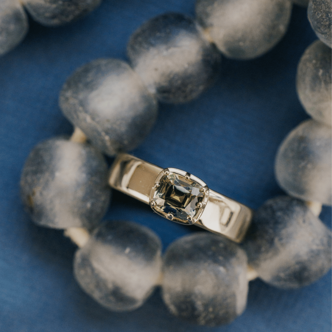 A 14k gold ring with a cushion-cut green Montana Sapphire set on a textured blue background on a beaded necklace.&nbsp;