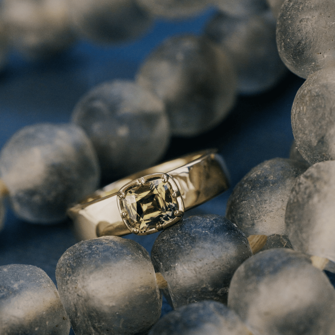 A 14k gold ring with a cushion-cut green Montana Sapphire set on a textured blue background on a beaded necklace.&nbsp;
