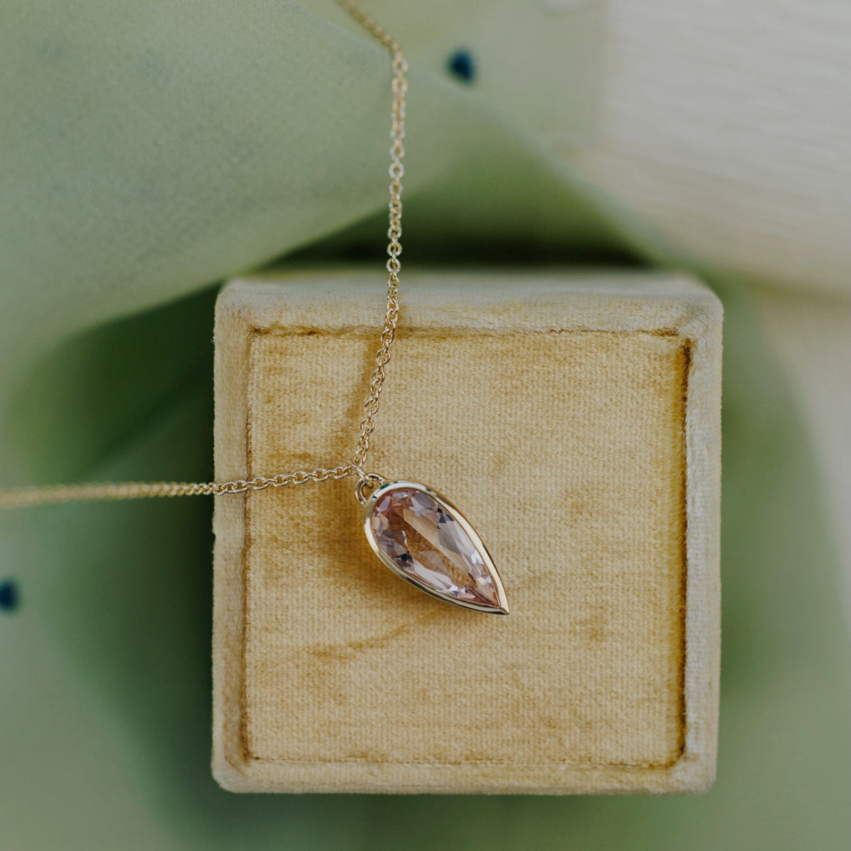 A 14k yellow gold pear-shaped morganite necklace in a bezel setting on a soft tan jewelry box with a textured background 
