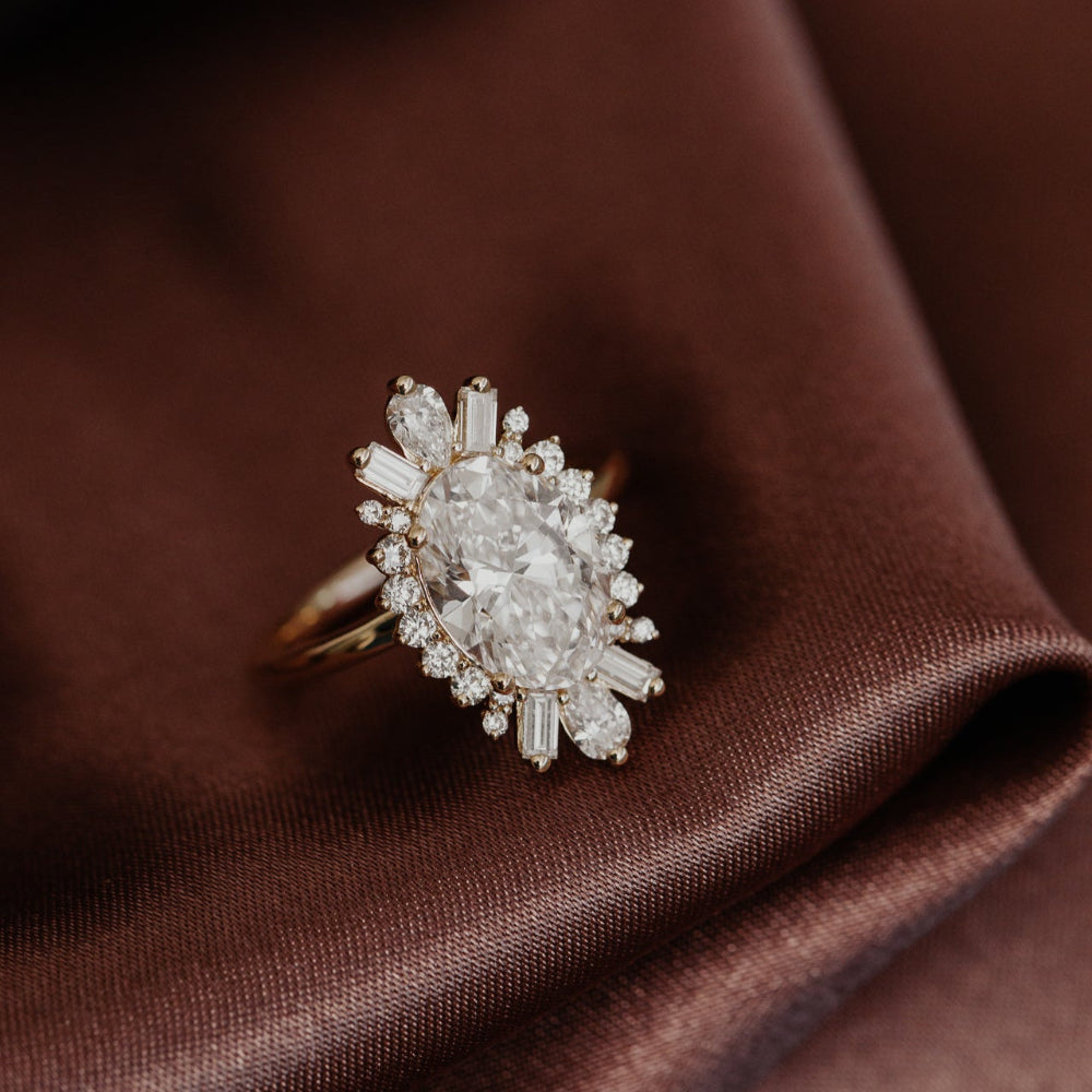 The Celeste Oval Diamond Ring with a halo design on a brown silk fabric. 
