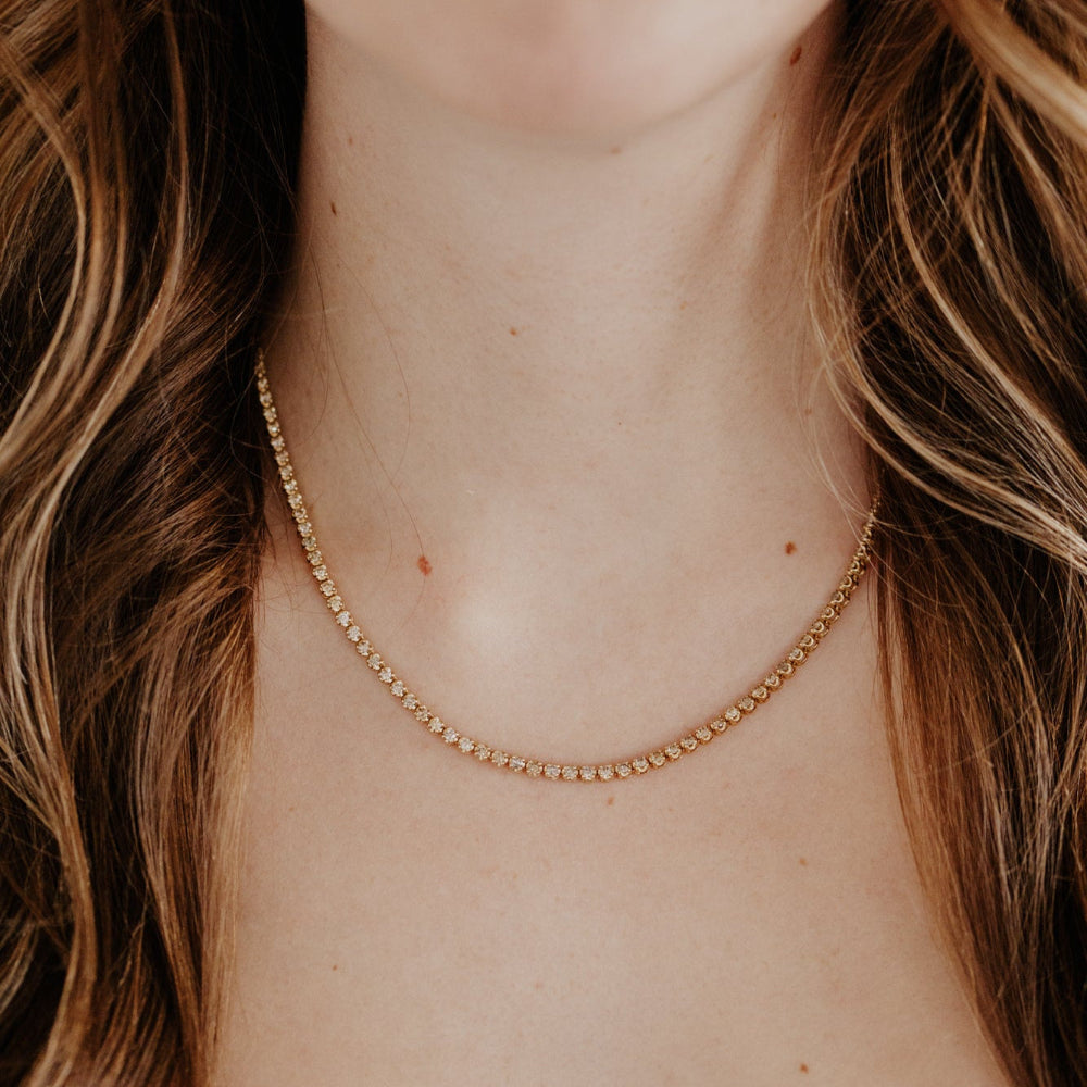 Close-up of a person wearing a gold champagne diamond tennis necklace with a blurred background.
