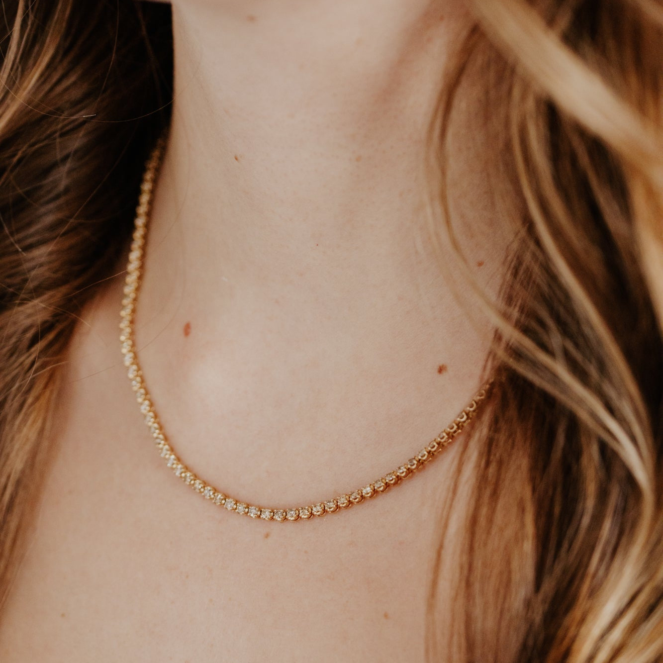 Close-up of a person wearing a delicate gold champagne diamond necklace with blurred hair in the foreground.