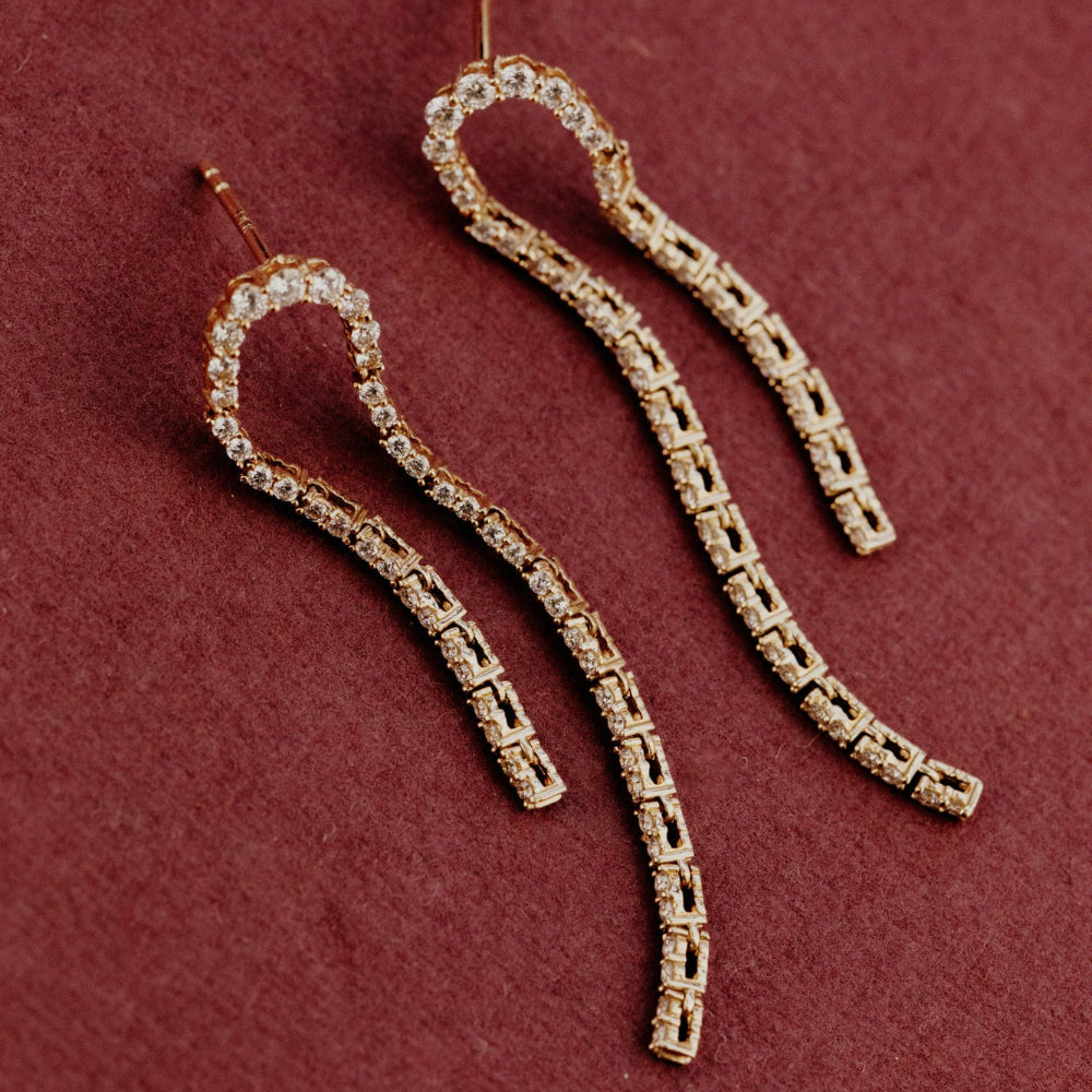 Gold curved diamond dangle earrings on a maroon background.